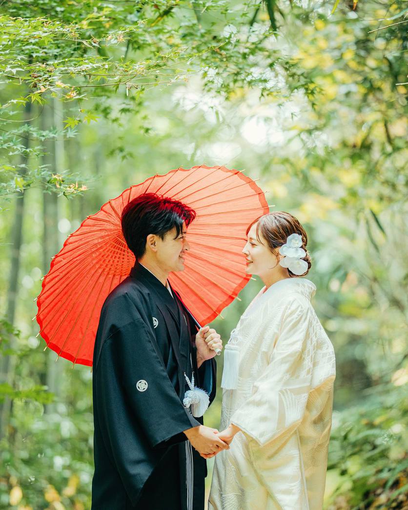 竹林で赤い番傘を差して向かい合う白無垢と黒紋付の二人